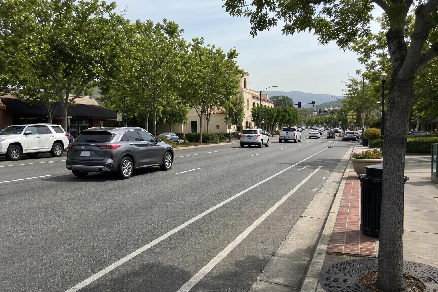 Wide tree-lined commercial boulevard with cars in several lanes, a bike lane, sidewalk with trash can, beige storefront buildings and hills in the distance