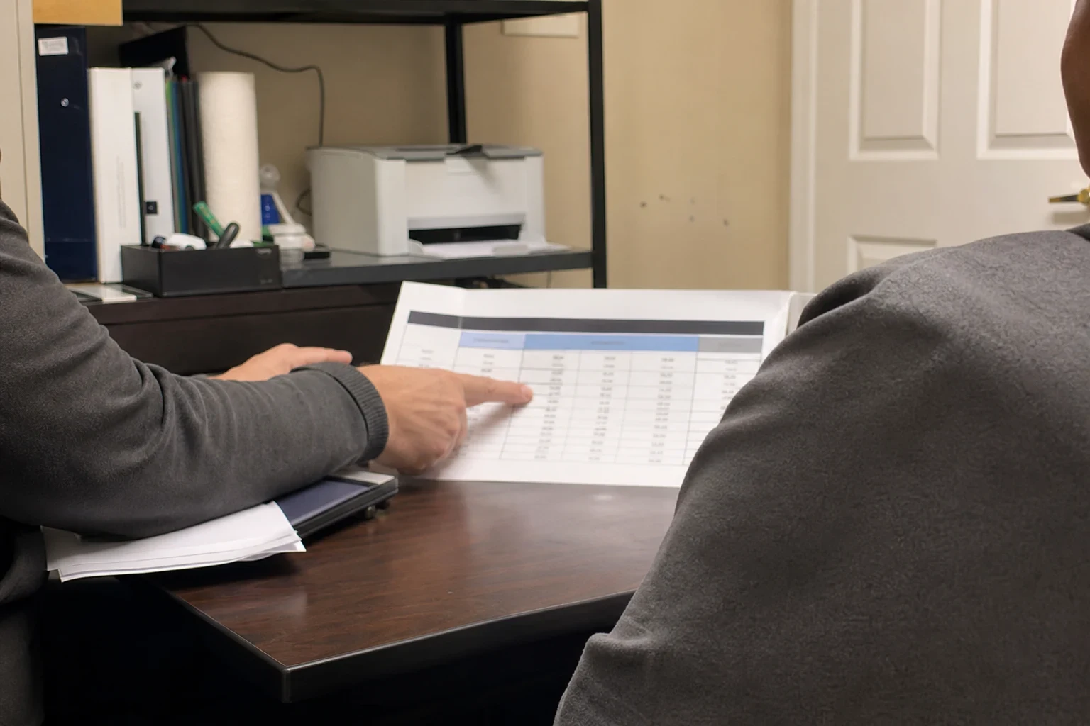 Agent's hand pointing to a printed table of coverage amounts on a dark wood desk in a small back office with a printer, binders and shelf behind