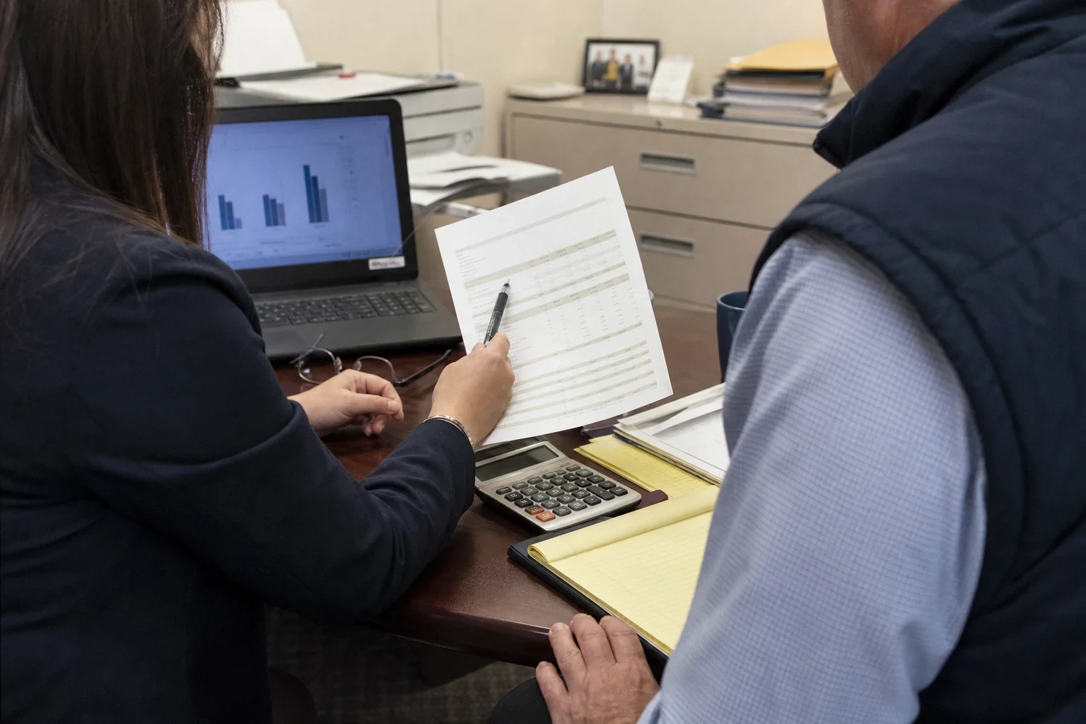 Over-the-shoulder view of a woman holding a statement and pen beside a man in a vest at an office desk with a laptop, calculator, yellow legal pad and filing cabinet.